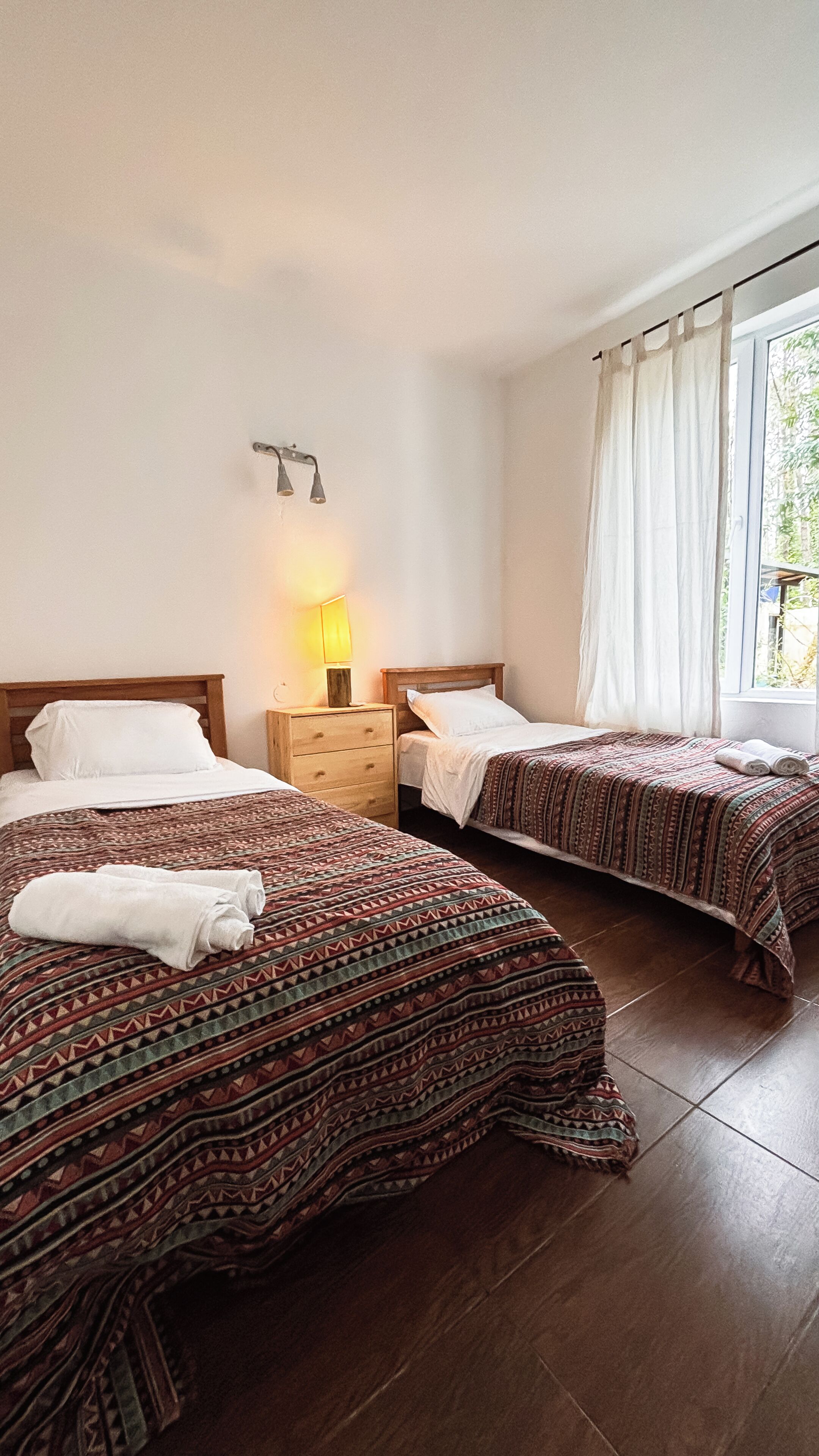 A hotel room with two beds, a bedside table with a lamp, and a window with curtains.