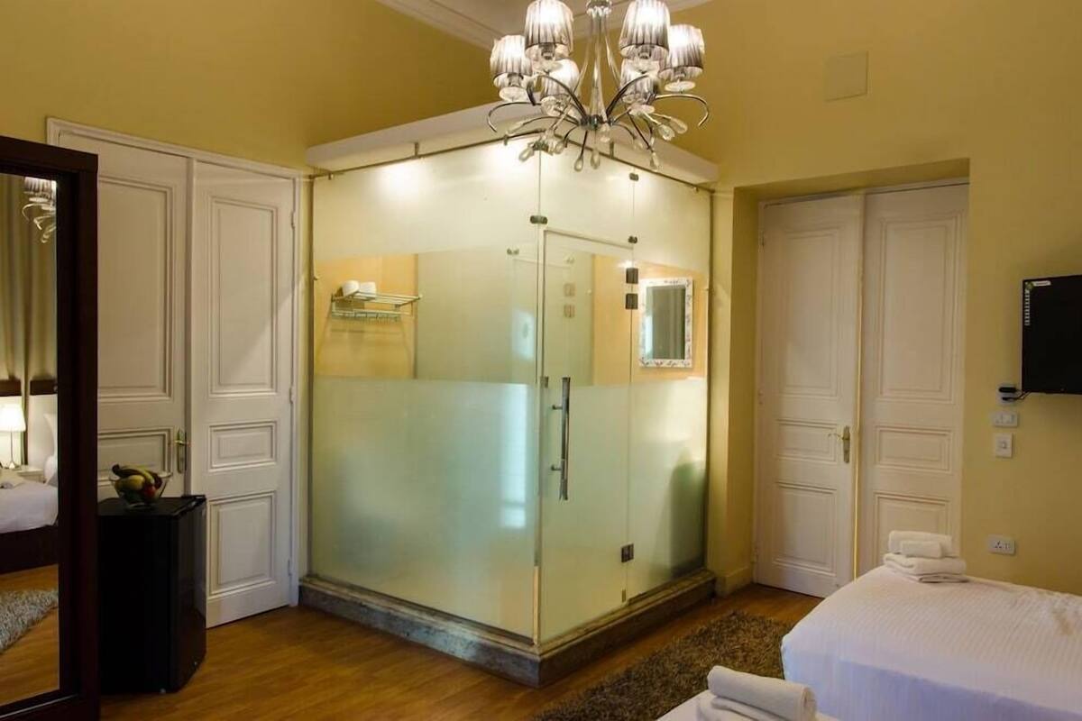 A hotel room with a walk-in closet, a bed with white linens, a chandelier, and a television mounted on the wall.