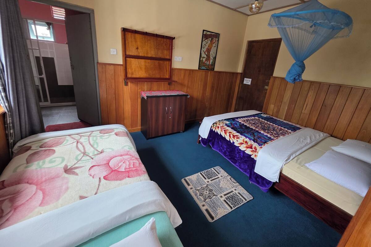 A hotel room with two beds, a wooden wardrobe, a small cabinet, a mosquito net, and a door leading to another room.