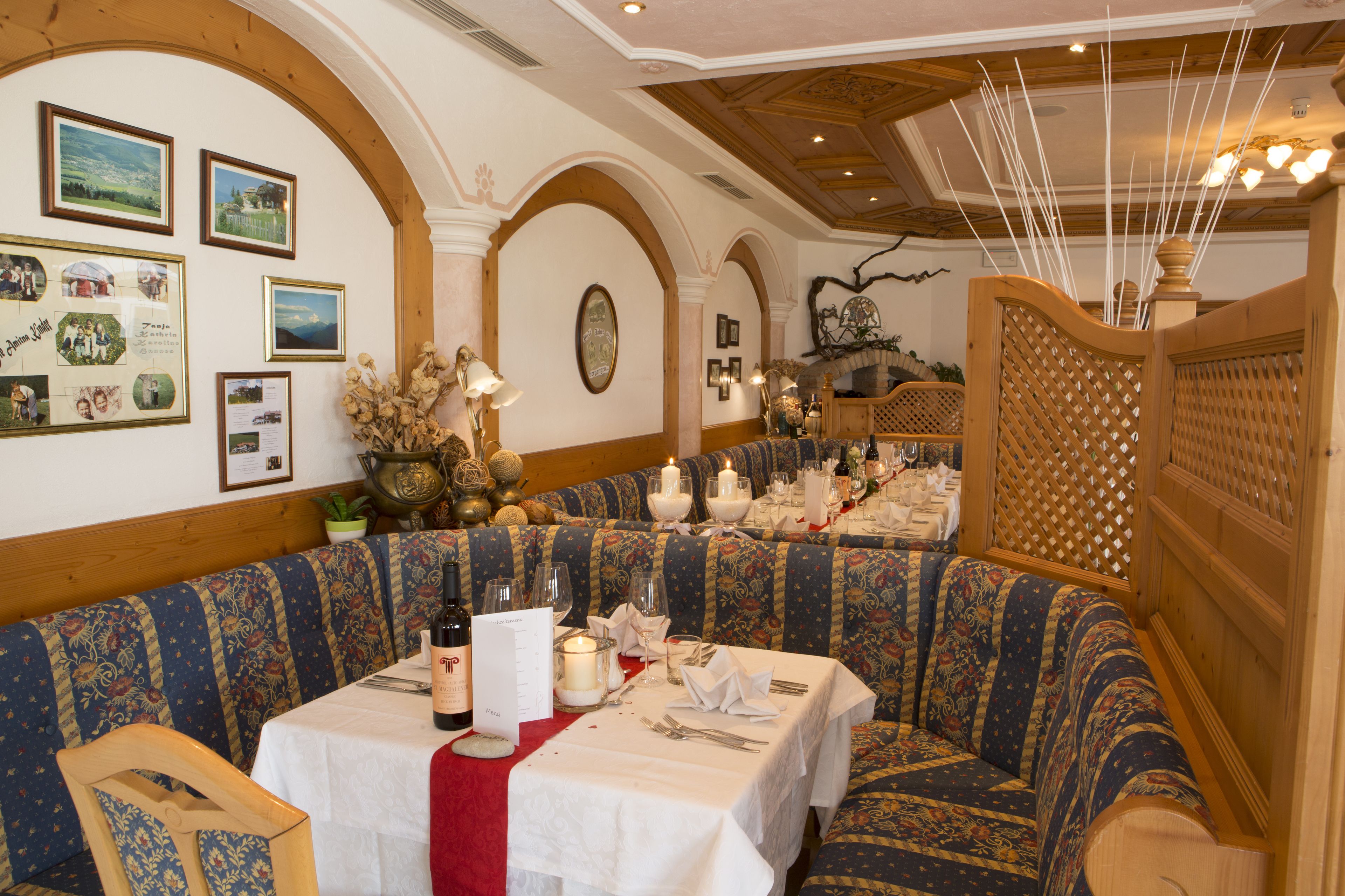 A restaurant with a cozy seating area featuring patterned blue and red upholstered benches. Tables are set with white tablecloths, wine glasses, and candles.