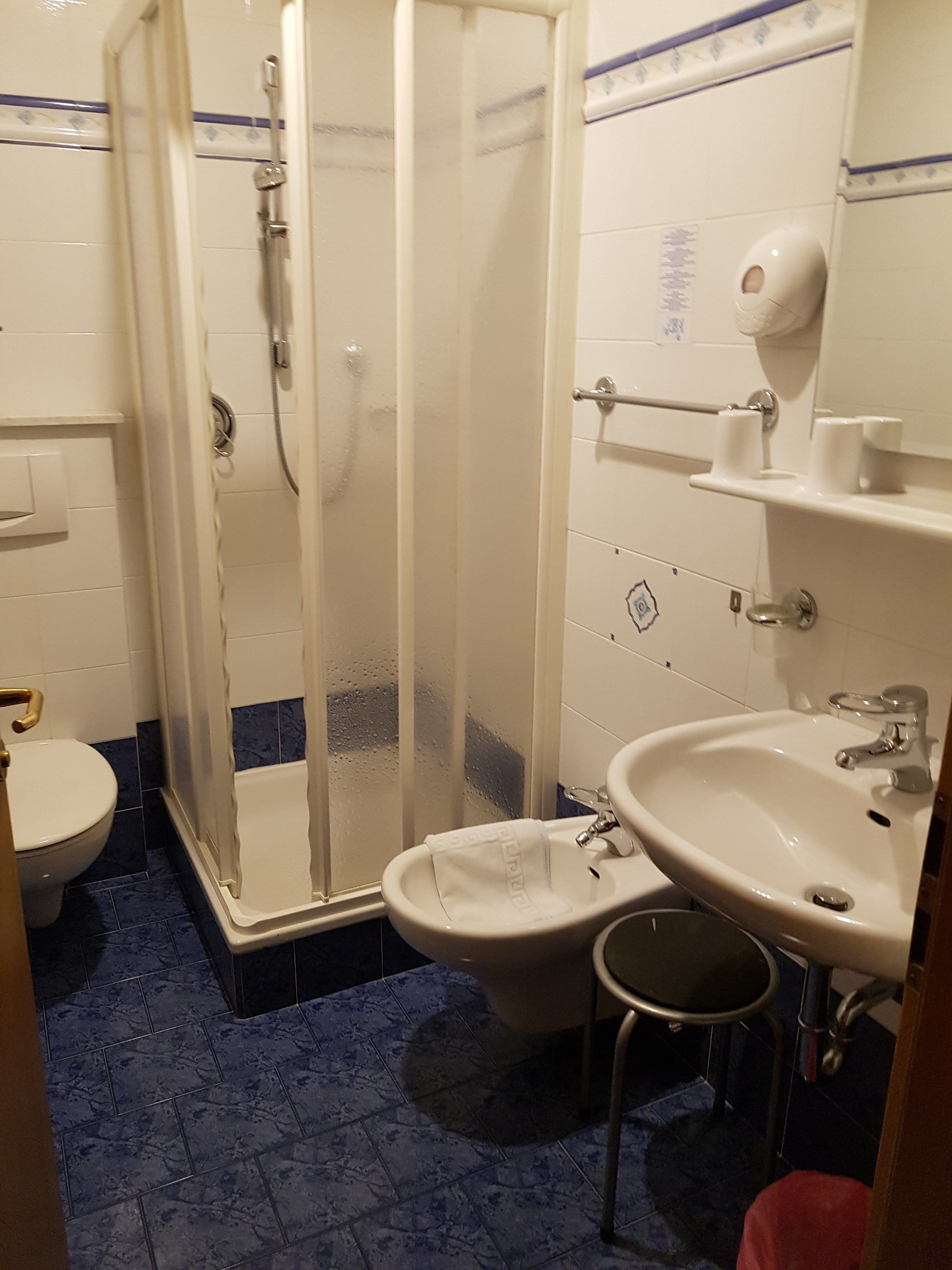Un baño con ducha, inodoro, lavamanos y un taburete pequeño.