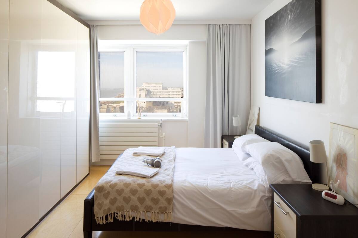 A hotel room with a bed, a bedside table, a lamp, and a view of the city through the window.