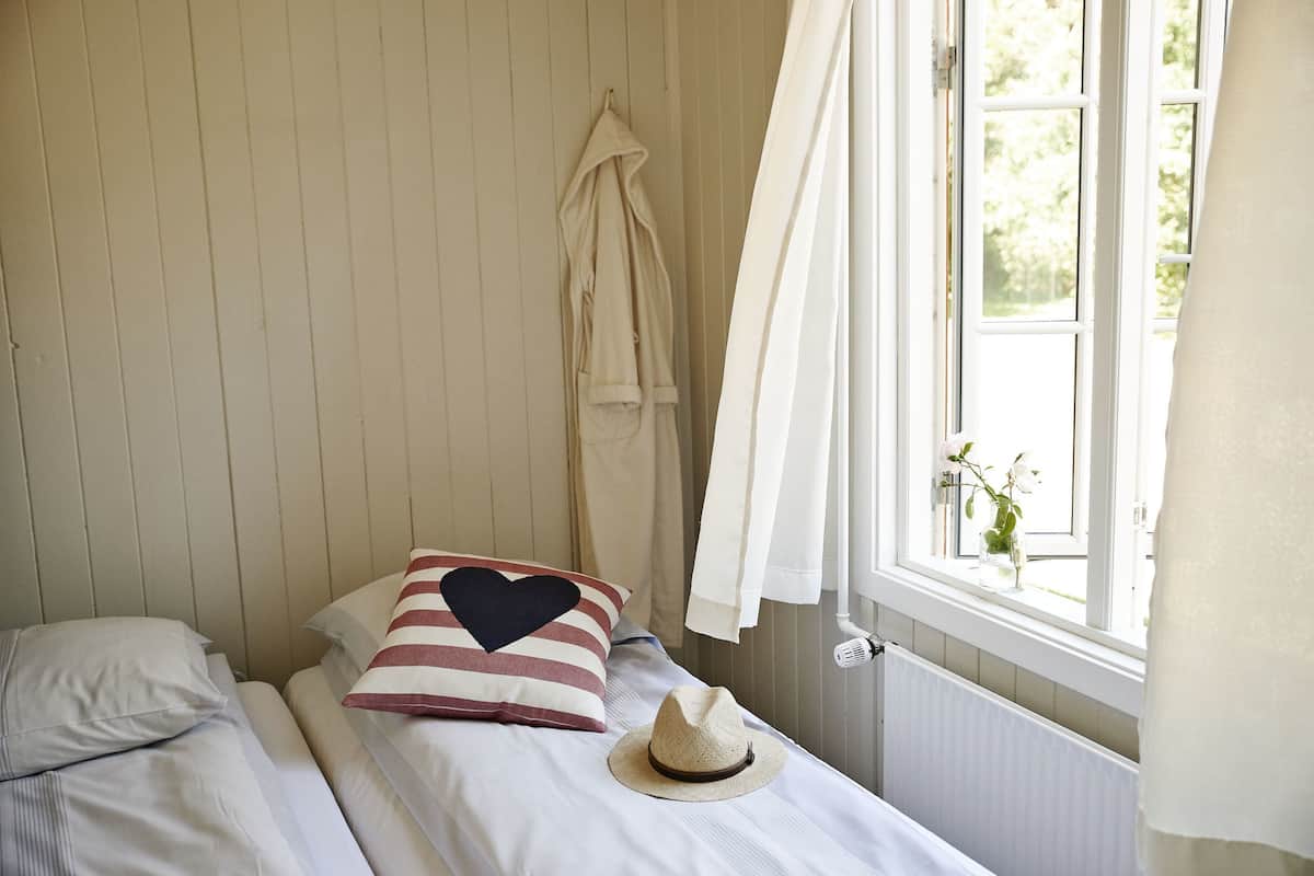 A bedroom with two beds, a window with curtains, a radiator, and a hat on one bed.