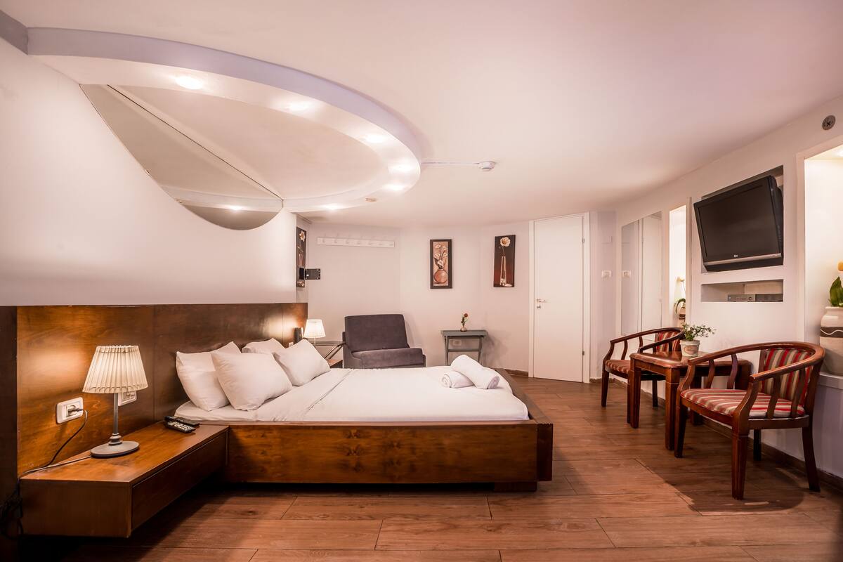A modern hotel room with a large bed, bedside table with lamp, a chair, a small table with a plant, and a TV mounted on the wall.