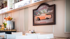 A restaurant interior with a vintage car poster, white chairs, and a wooden table. - Avital Hotel (Jerusalem)