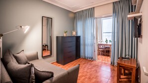 A living room with a sofa, a wooden table, a mirror, a dresser, and a view of the outdoors through a window with curtains. - Avital Hotel (Jerusalem)