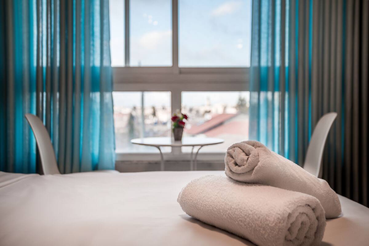 A hotel room with a bed, two rolled towels, a small table with a vase, and a view through a window with curtains.