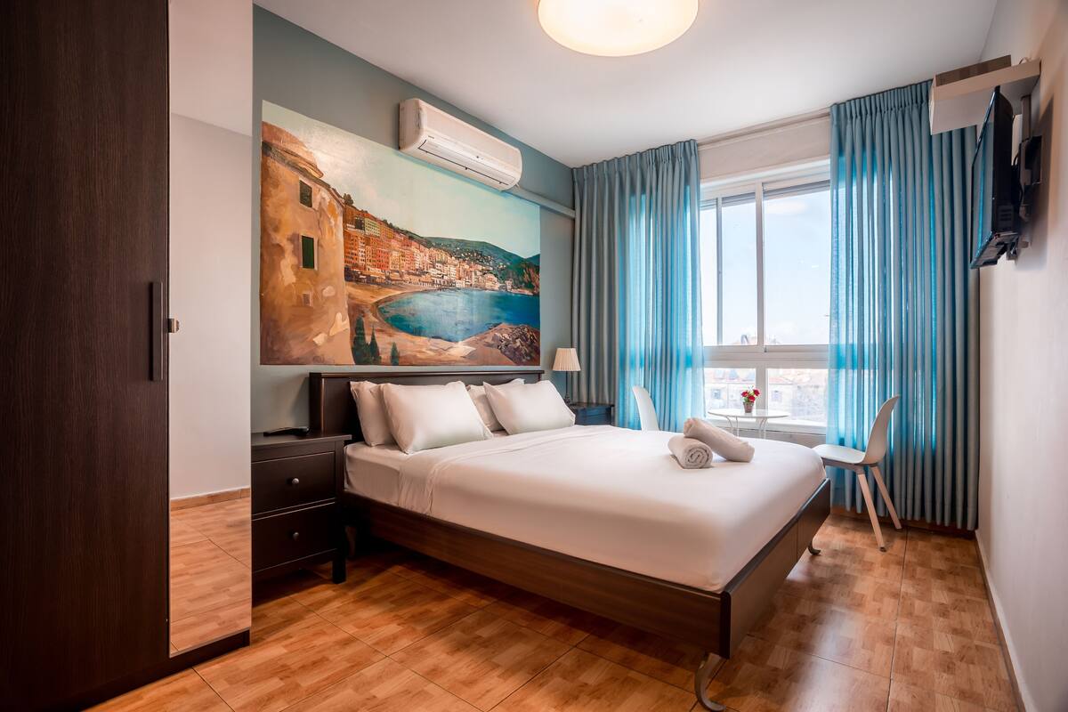 A hotel room with a bed, a desk, a chair, a window with curtains, and a painting on the wall.