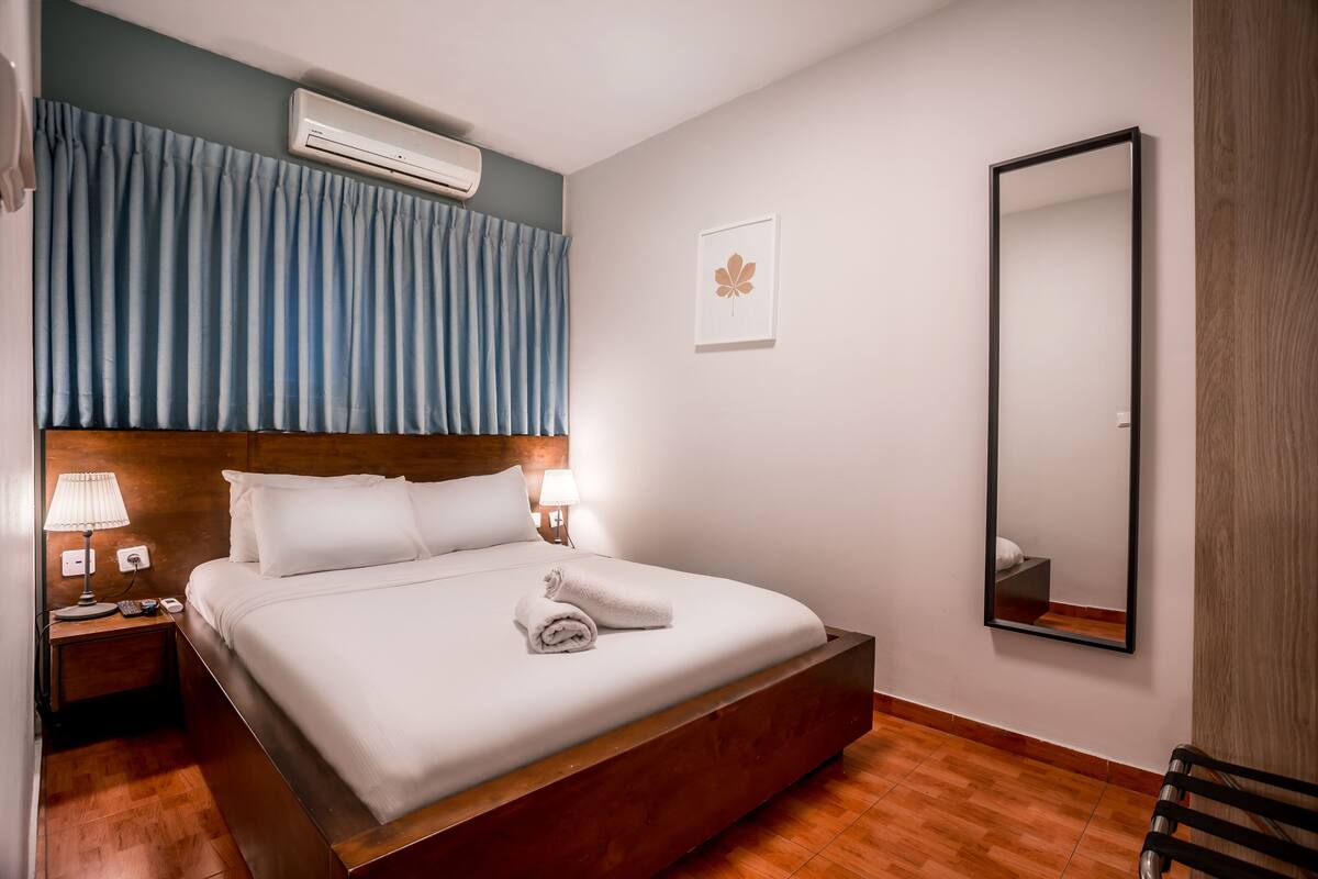 A hotel room with a large bed, a nightstand with a lamp, a wall-mounted air conditioner, a framed picture, and a mirror.