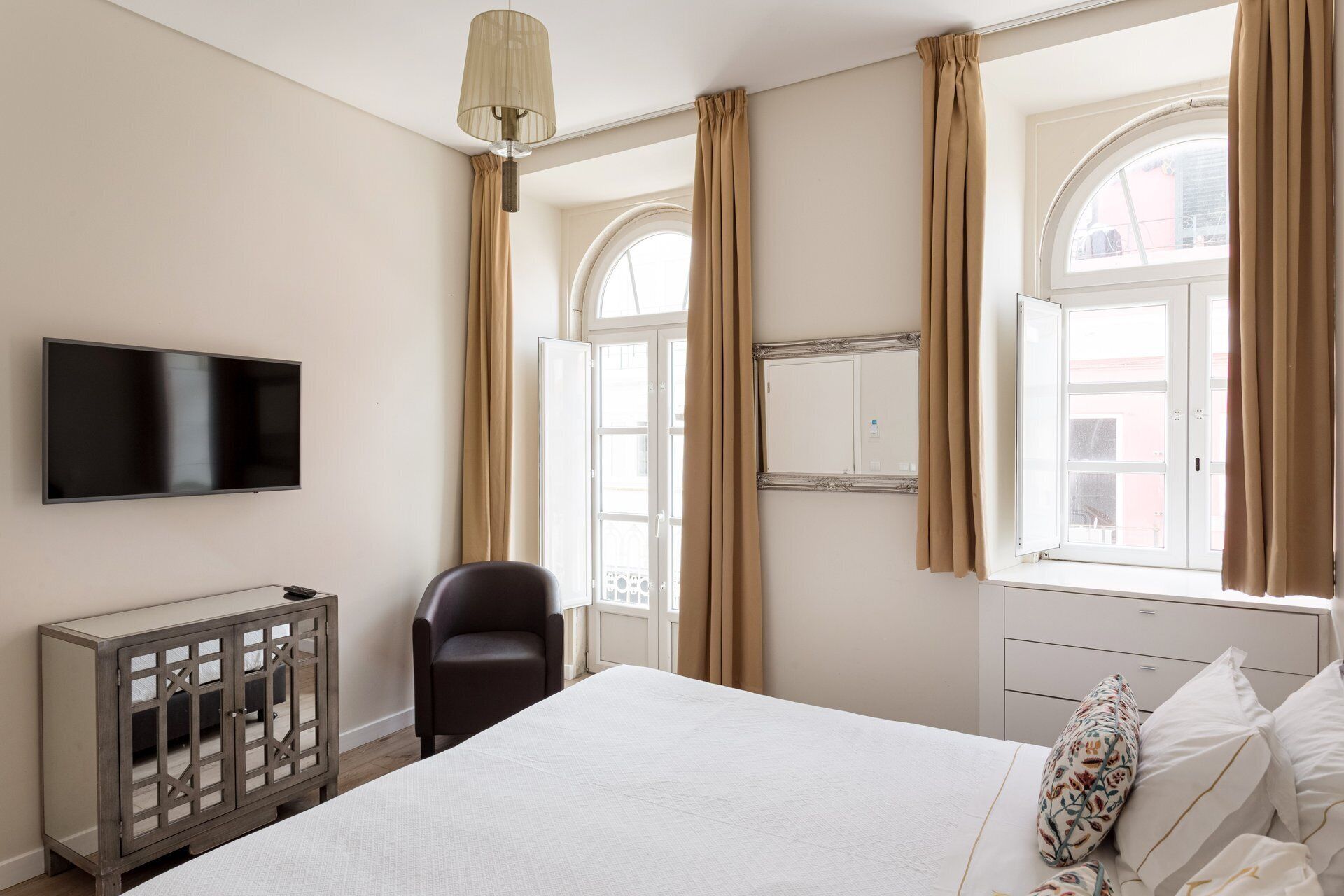 A bedroom with a bed, a TV, a chair, a cabinet, and large windows with curtains.