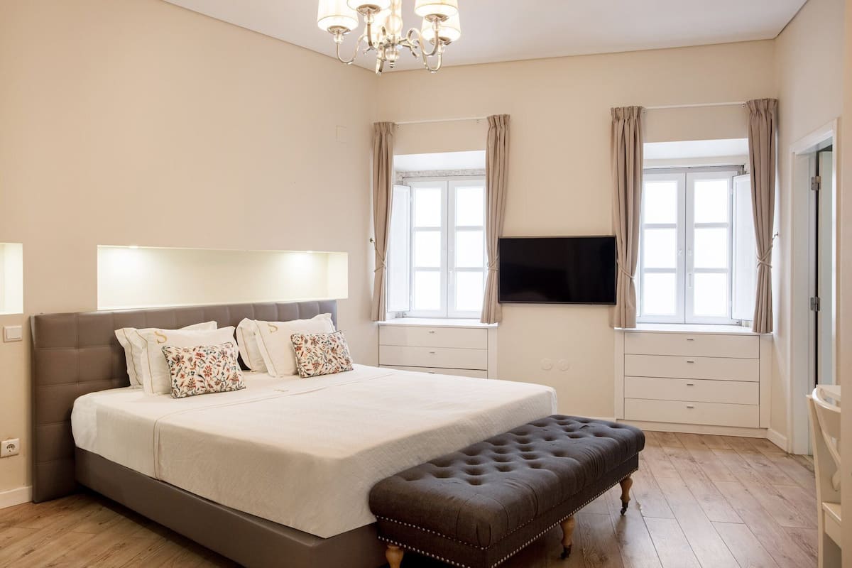 A bedroom with a large bed, a bench, a TV, and two windows with curtains.