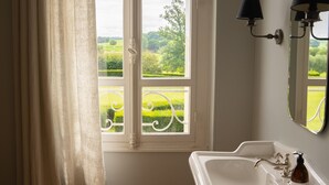 A bathroom with a white sink, a window with a view of greenery, and a mirror with wall-mounted lights. - Chambre D'hôtes Château de Lussac (Lussac)