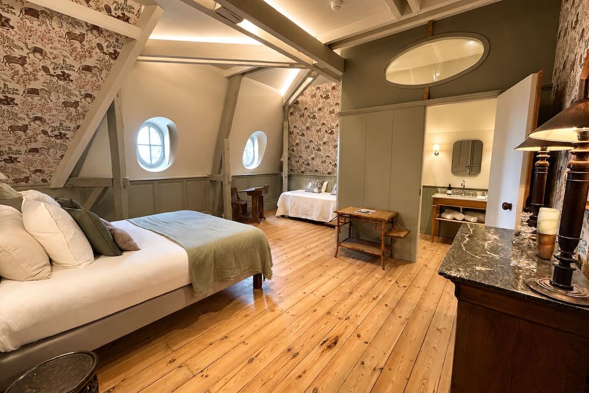 A bedroom with a bed, two windows, a small table, a mirror, and a bathroom with a sink and mirror.