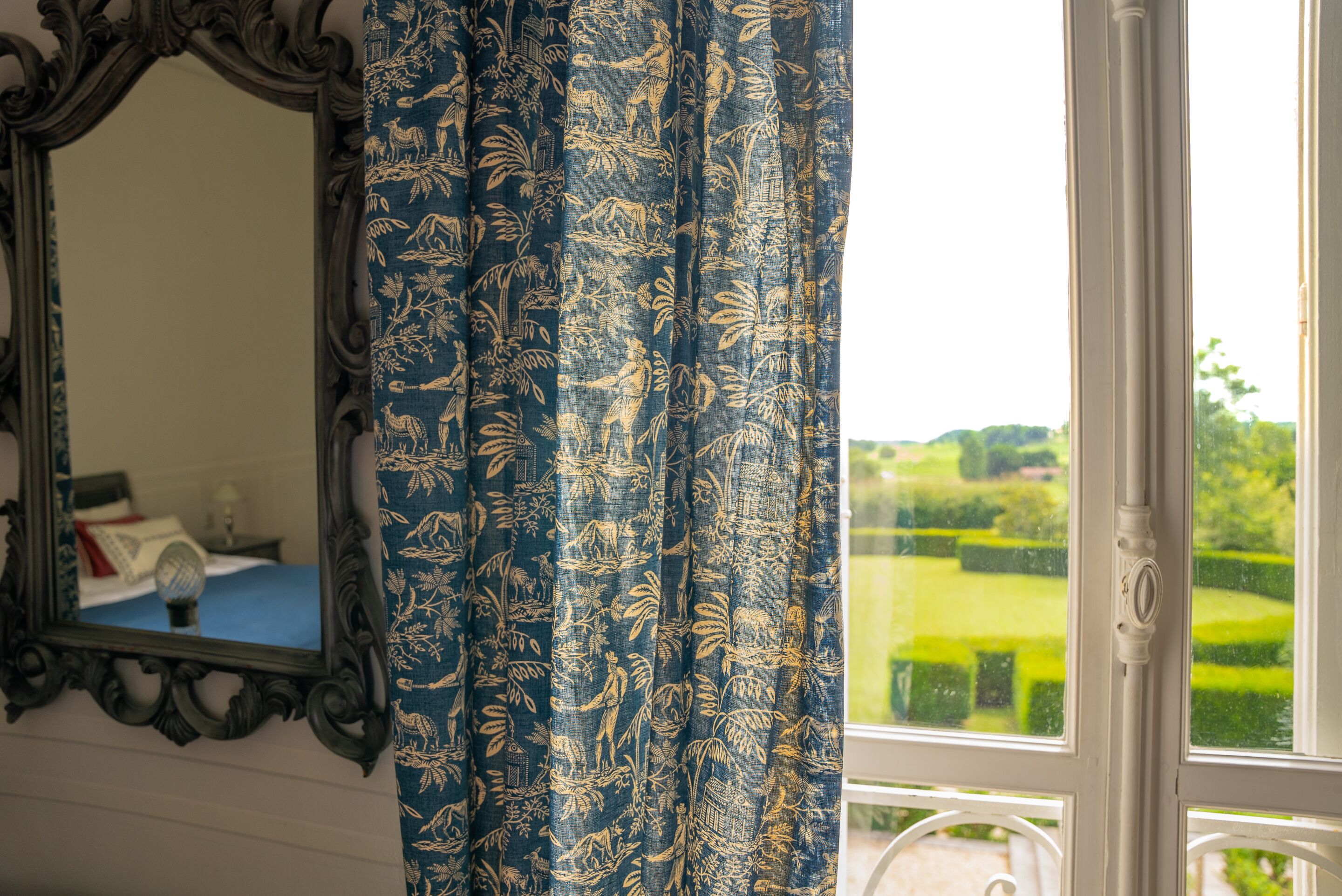 Una habitación con un espejo grande, una ventana con vista a un jardín cuidado y una cortina estampada.