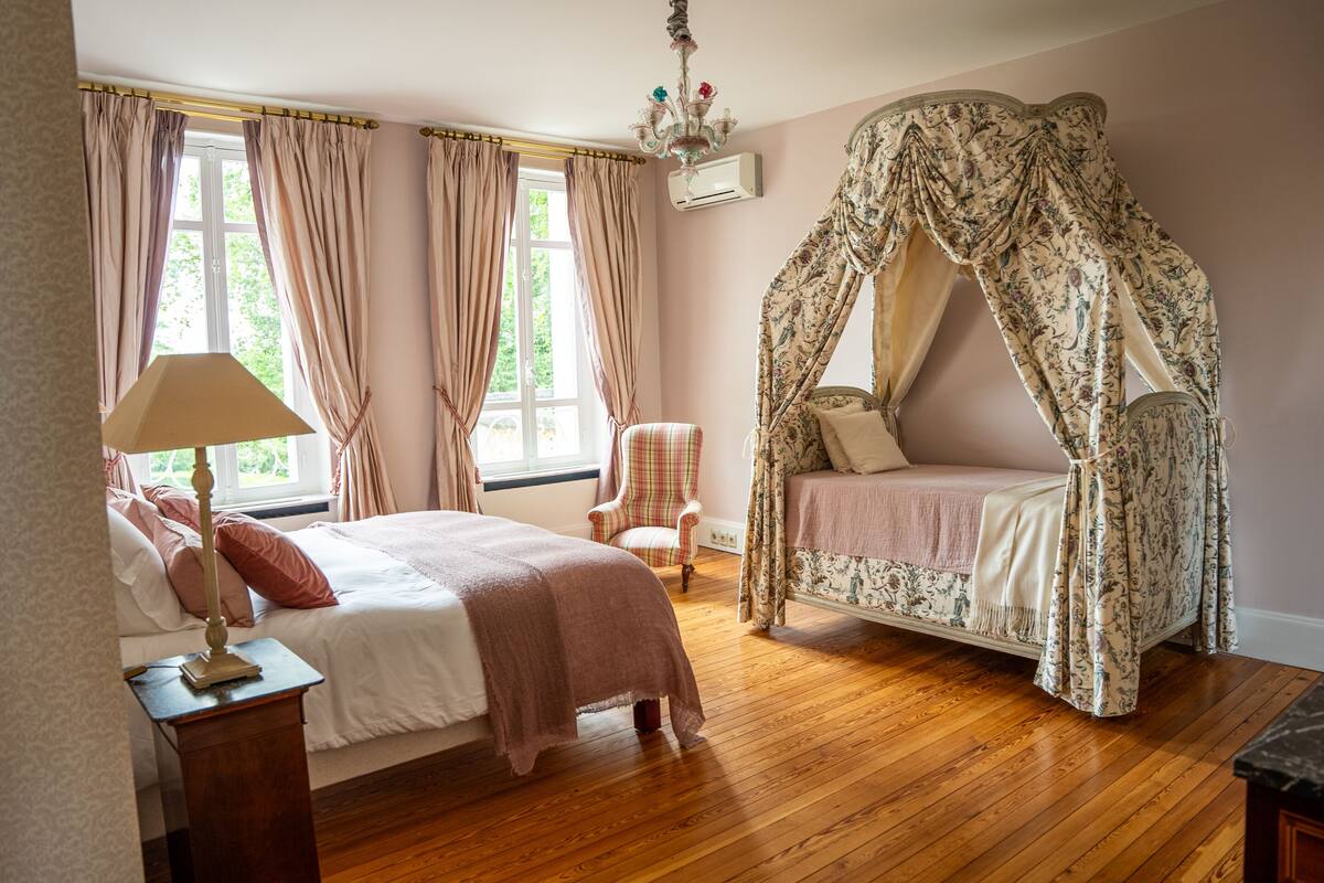 A bedroom with a four-poster canopy bed, a bedside table with a lamp, a chair, and a window with curtains.