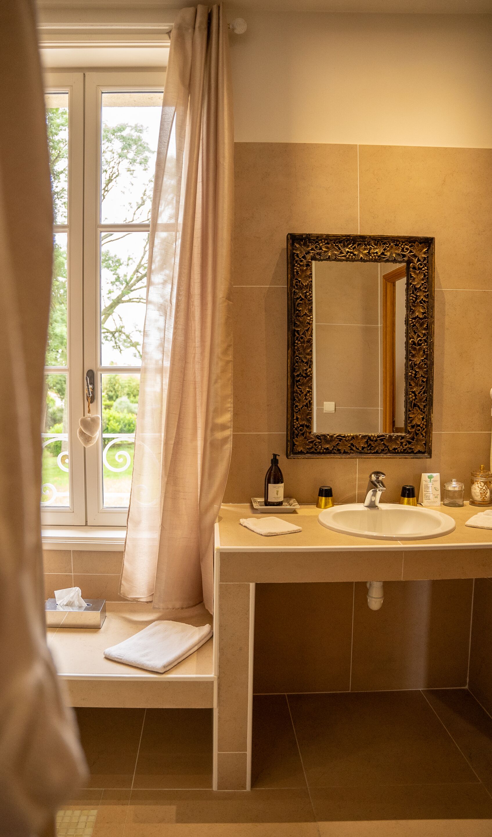 A bathroom with a large window, a mirror with an ornate frame, a sink, and a towel rack.