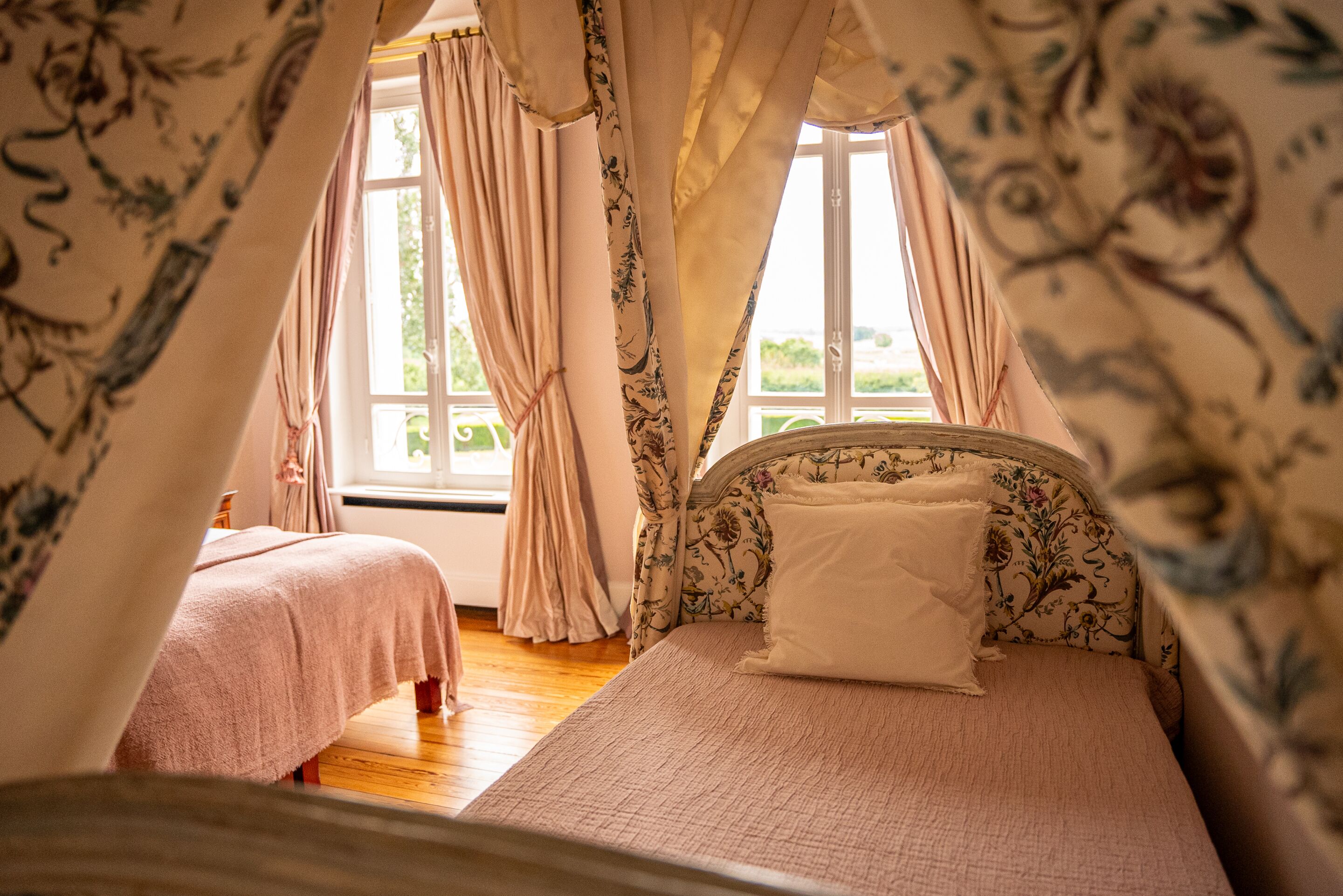Un dormitorio con dos camas, cortinas con estampado floral y vista a zonas verdes.