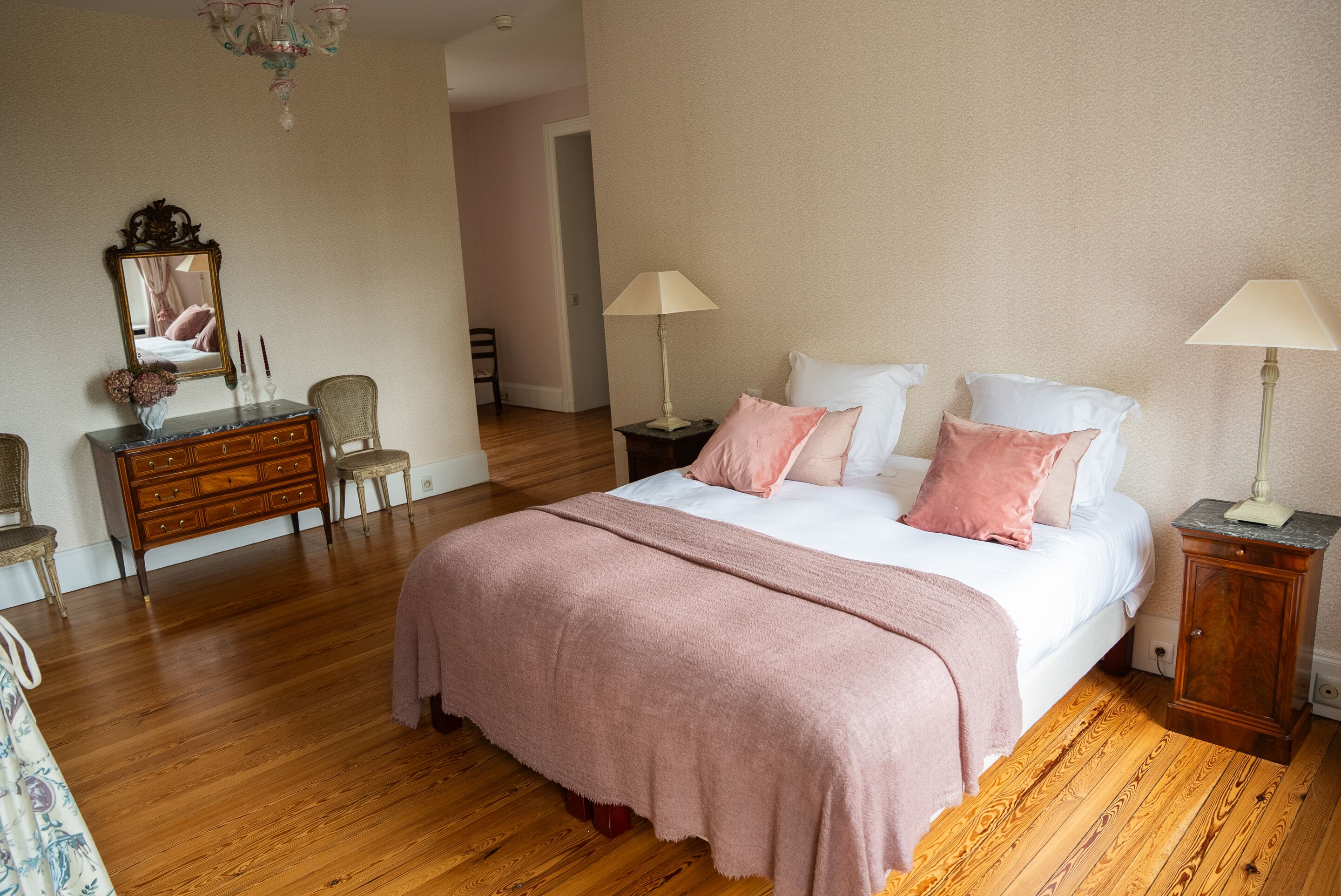 A bedroom with a large bed, wooden floor, a dresser, a mirror, and two bedside tables with lamps.