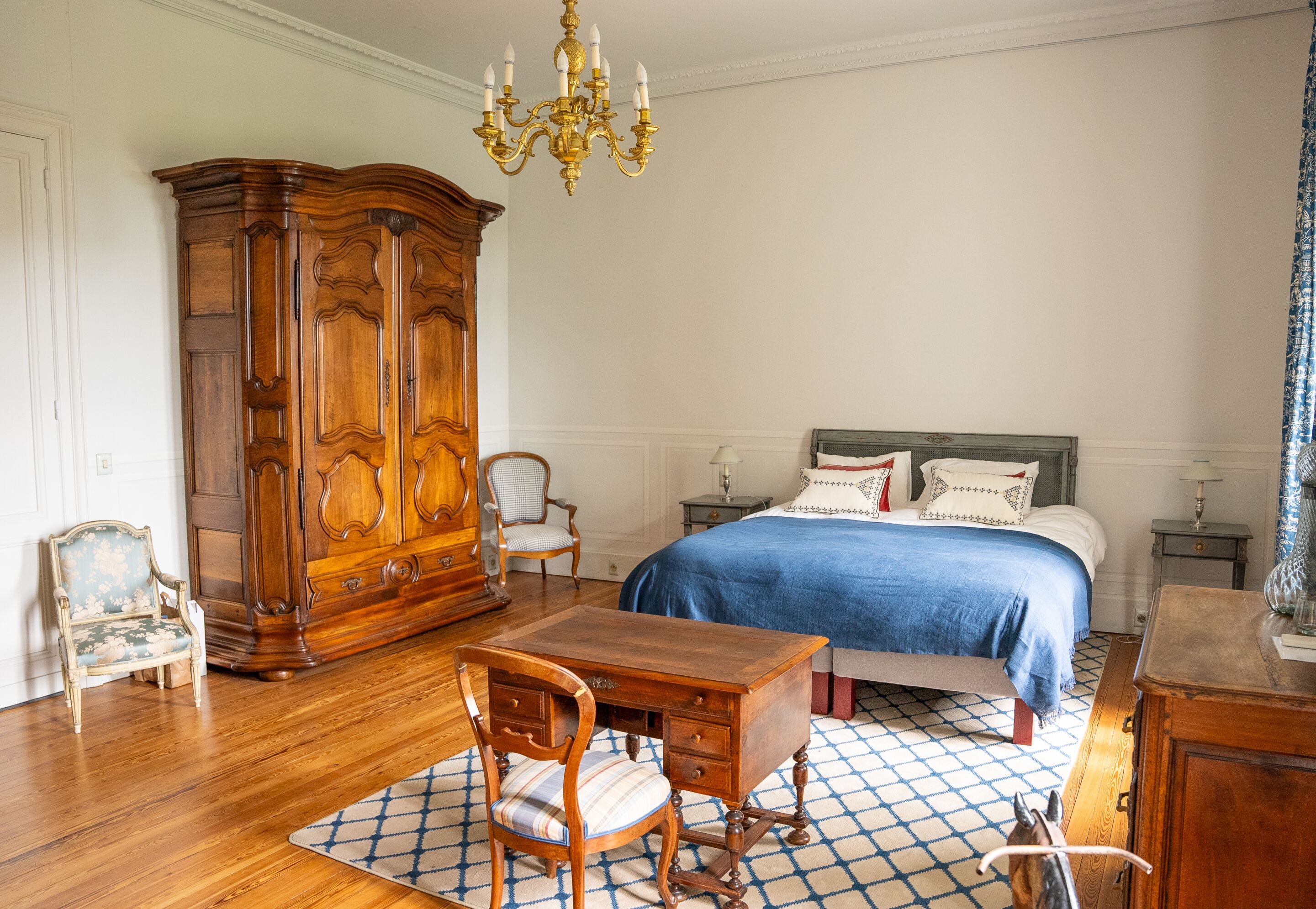 Une chambre avec une grande armoire en bois, un lit recouvert d’une courtepointe bleue, un bureau en bois avec une chaise et un lustre.