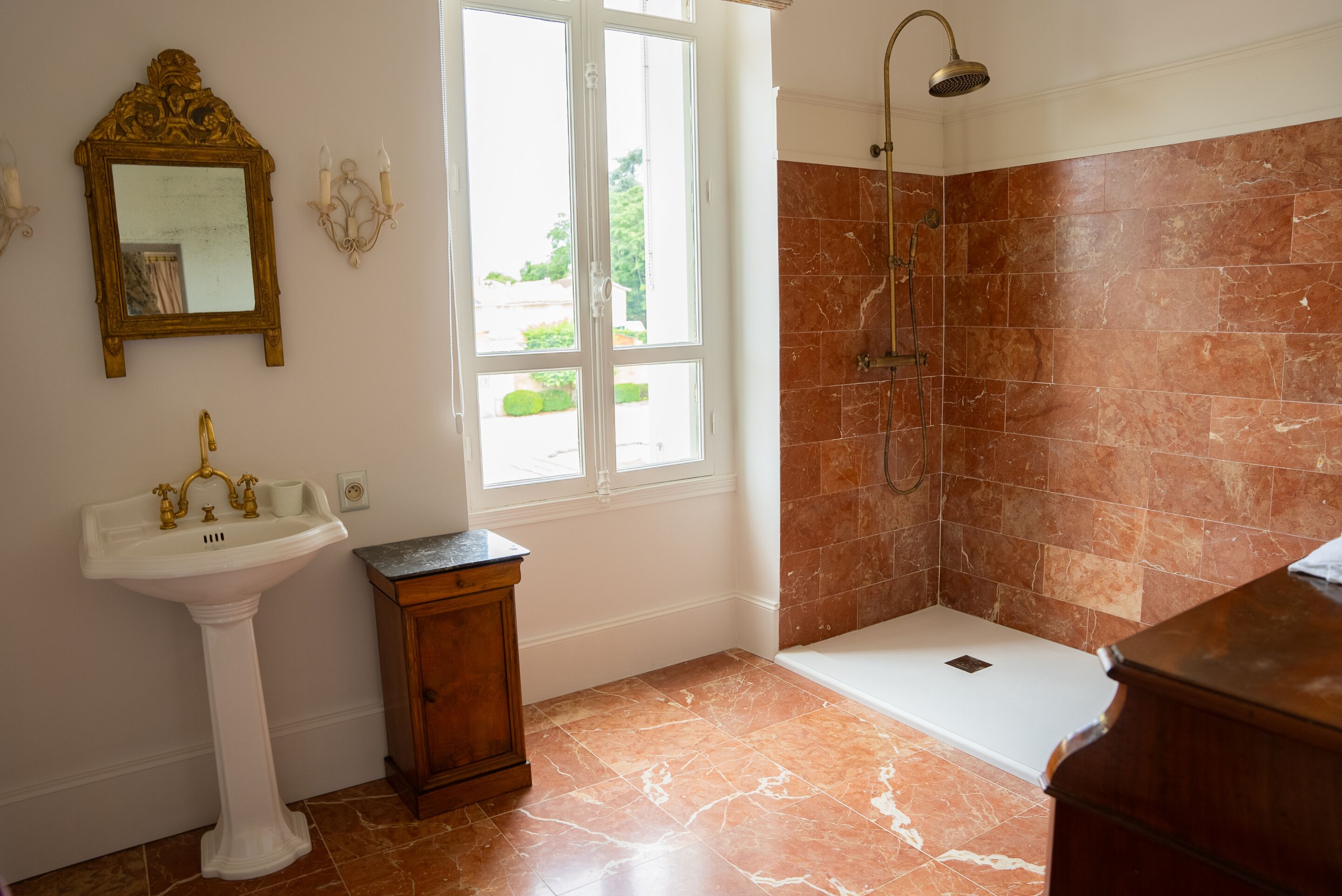 Een badkamer met een wastafel op een voetstuk, een douche met een handdouche en een raam dat voor natuurlijk licht zorgt.