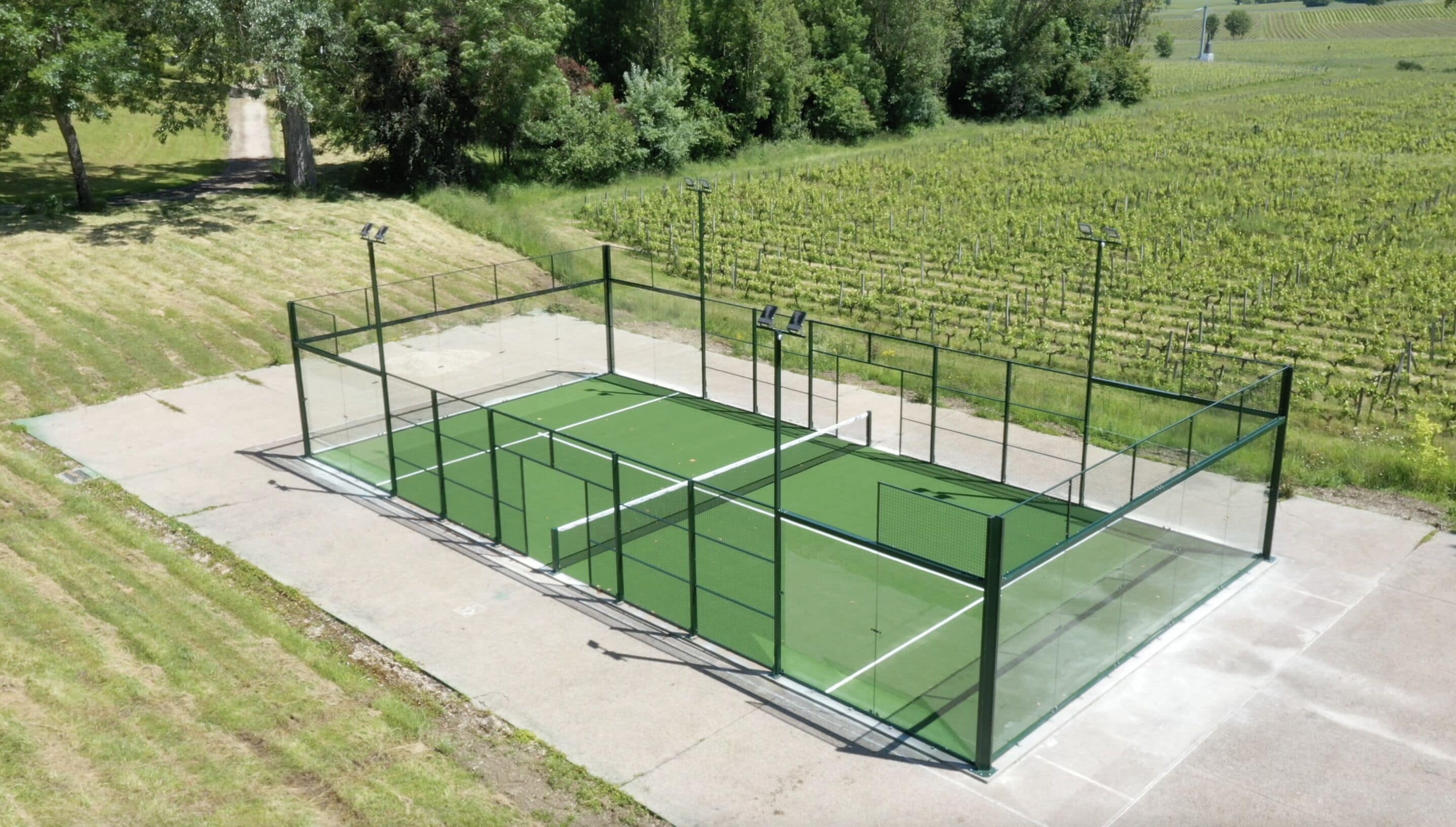 Una cancha de tenis con superficie verde, rodeada por una valla, ubicada en una zona con césped, árboles y un viñedo al fondo.