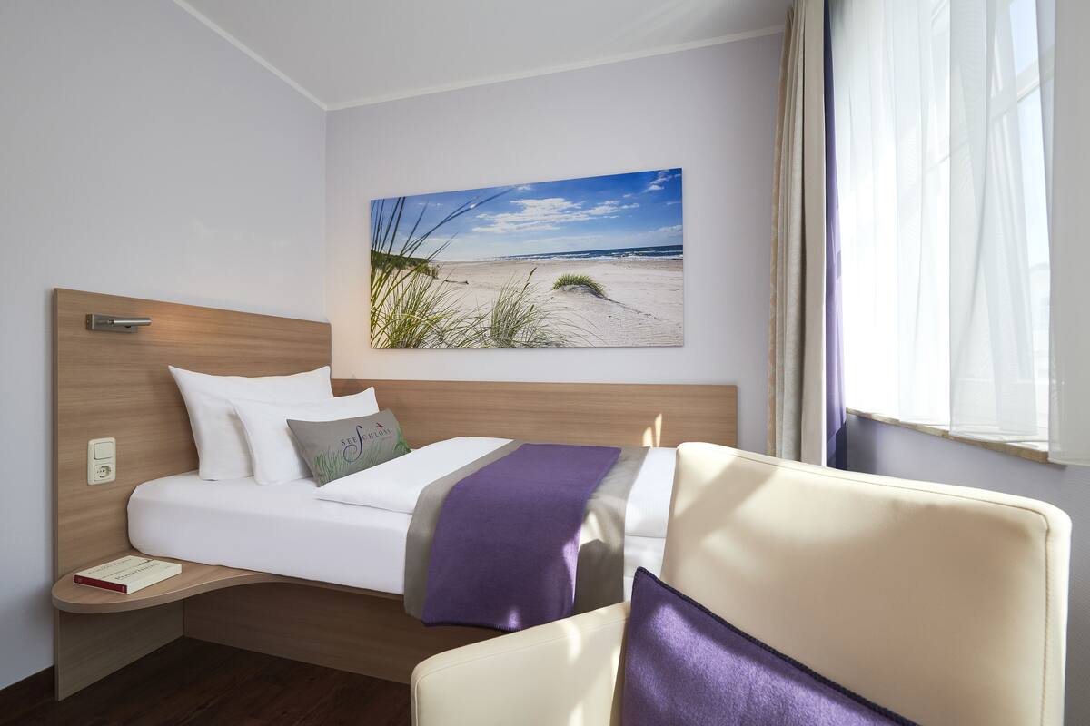 A hotel room with a bed, a chair, a nightstand, and a wall-mounted picture of a beach.