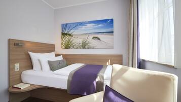 A hotel room with a bed, a chair, a nightstand, and a wall-mounted picture of a beach.