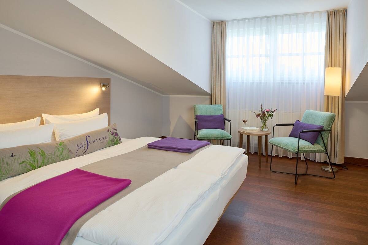 A hotel room with a large bed, two chairs, a small table, and a window with curtains.