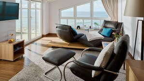 Uma sala de estar moderna com uma televisão grande, assentos confortáveis, uma mesa de centro de madeira e vista para o mar através de janelas amplas.