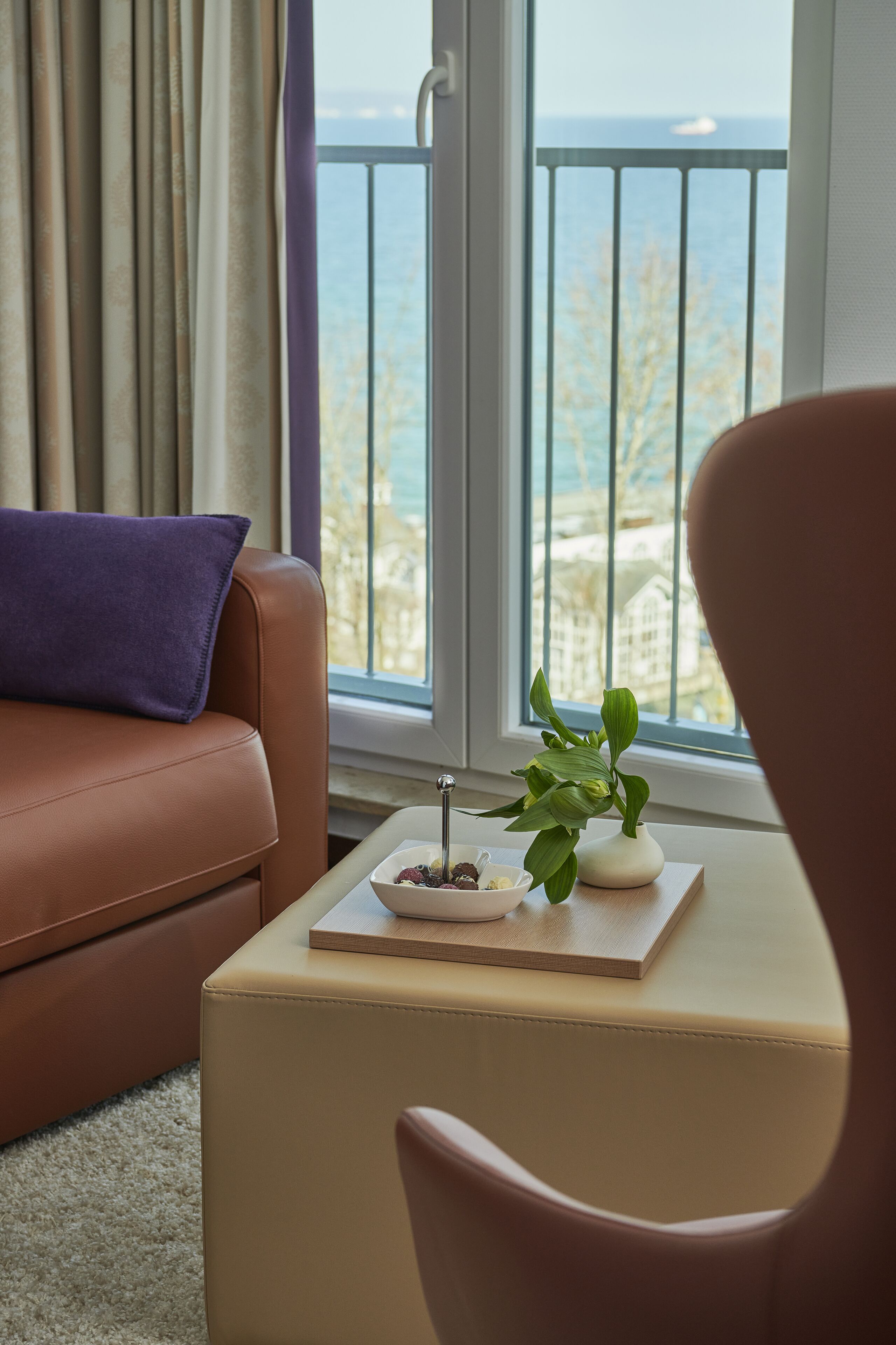 A living room with a brown leather sofa, a small table with a plant and a bowl of fruit, and a view of the sea through a balcony railing.