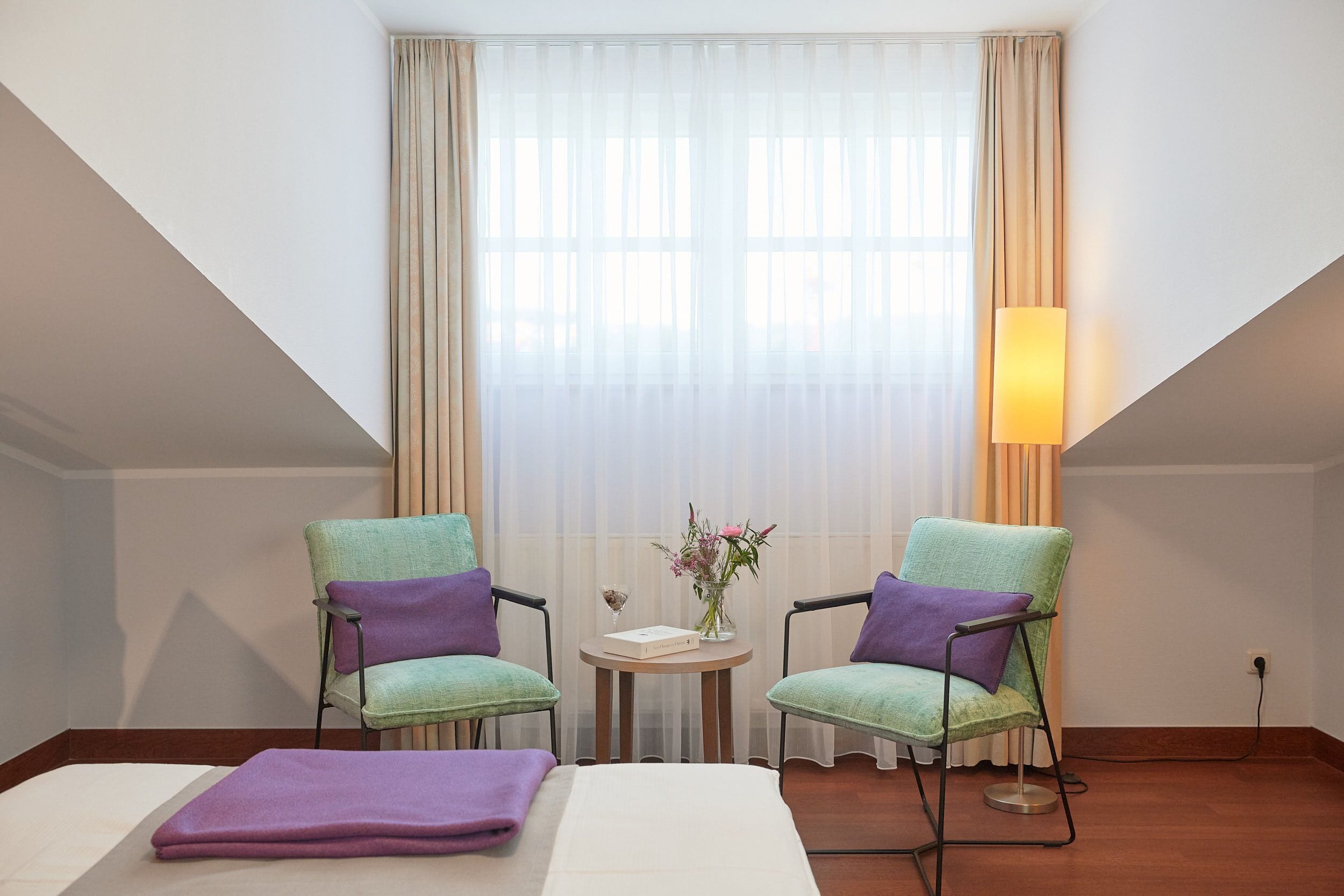 A hotel room with a bed, two chairs, a small table with a vase of flowers, and a lamp.