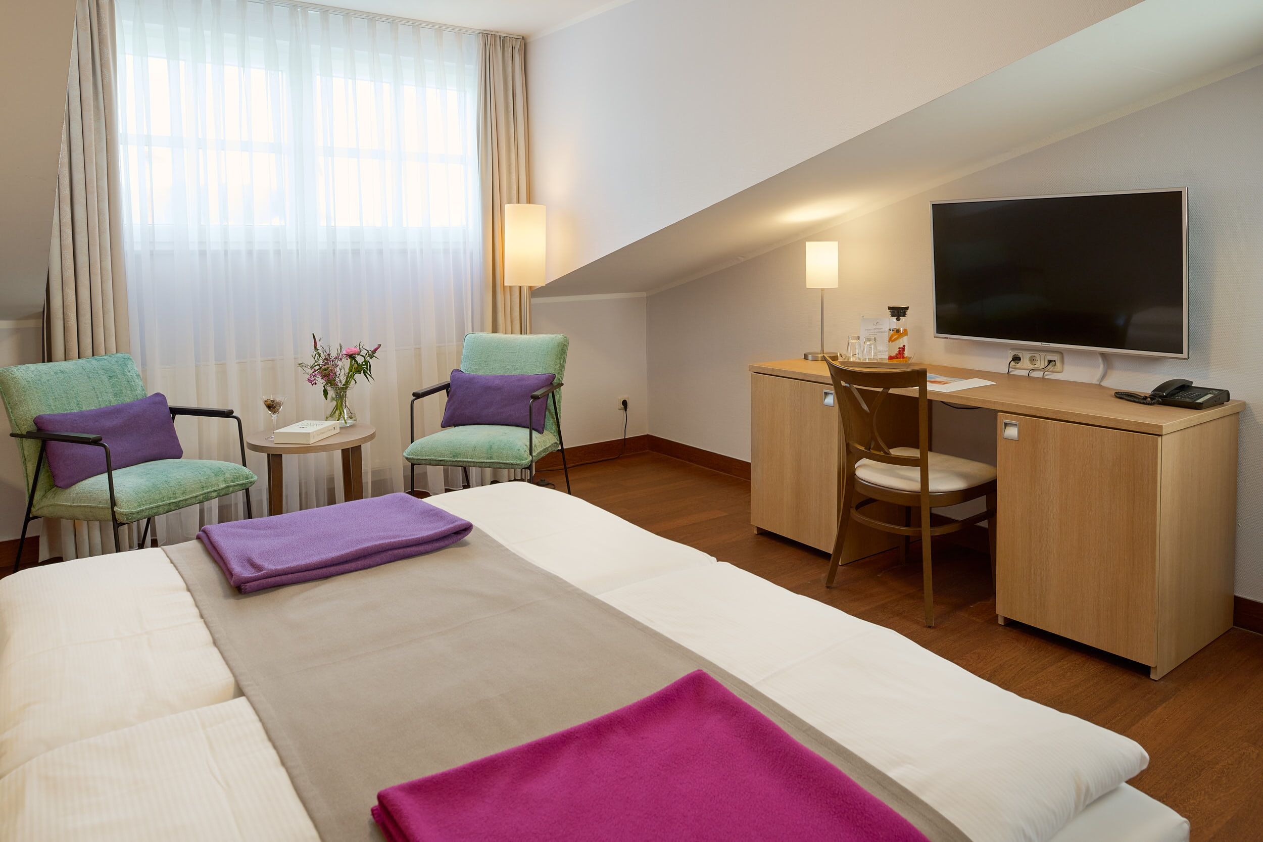 Ett hotellrum med en säng, två stolar, ett skrivbord, en tv och ett litet bord med en vas med blommor.