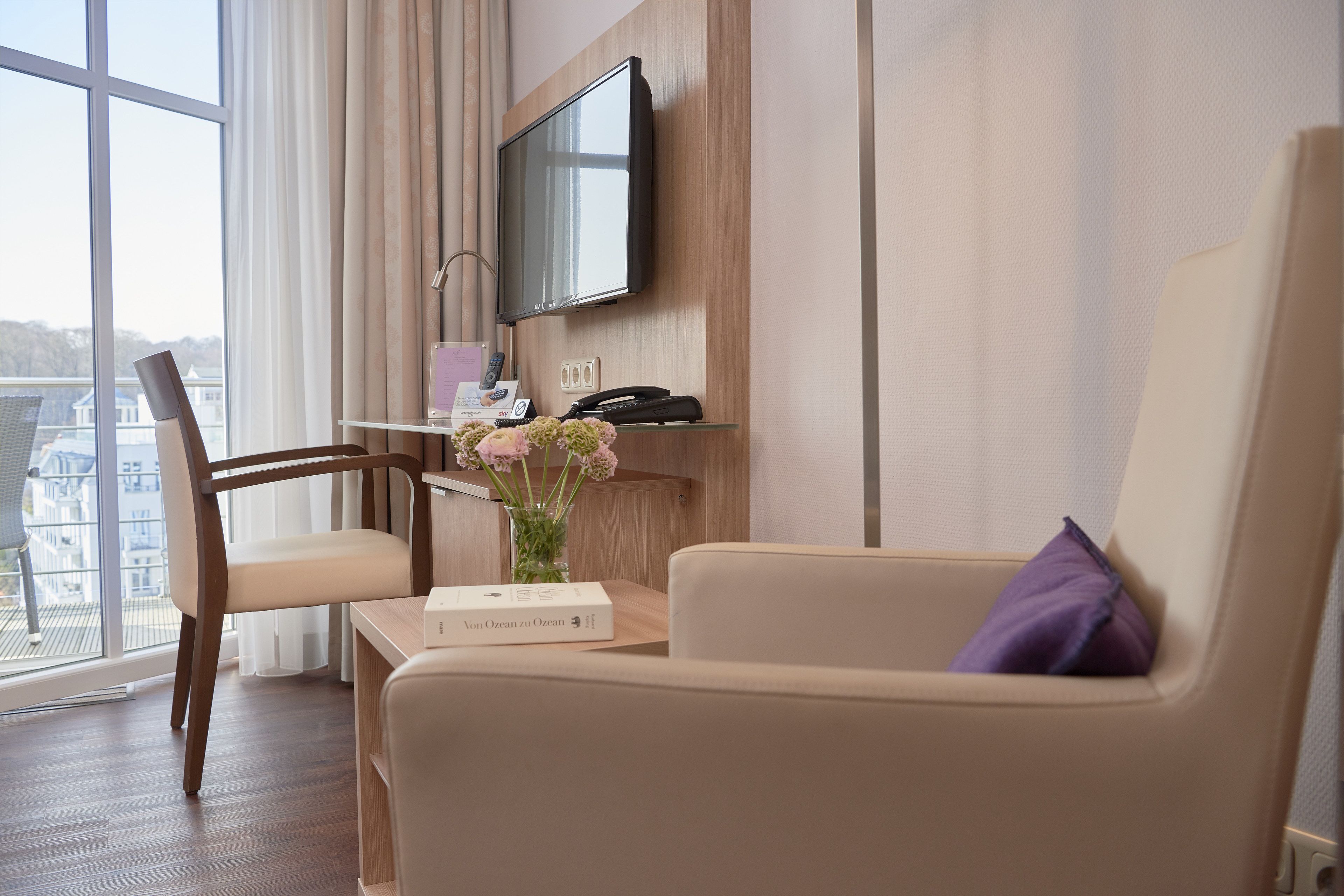 Quarto de hotel moderno com uma janela grande, uma TV de tela plana na parede, uma escrivaninha de madeira com telefone e um vaso de flores, e um sofá bege com uma almofada roxa.