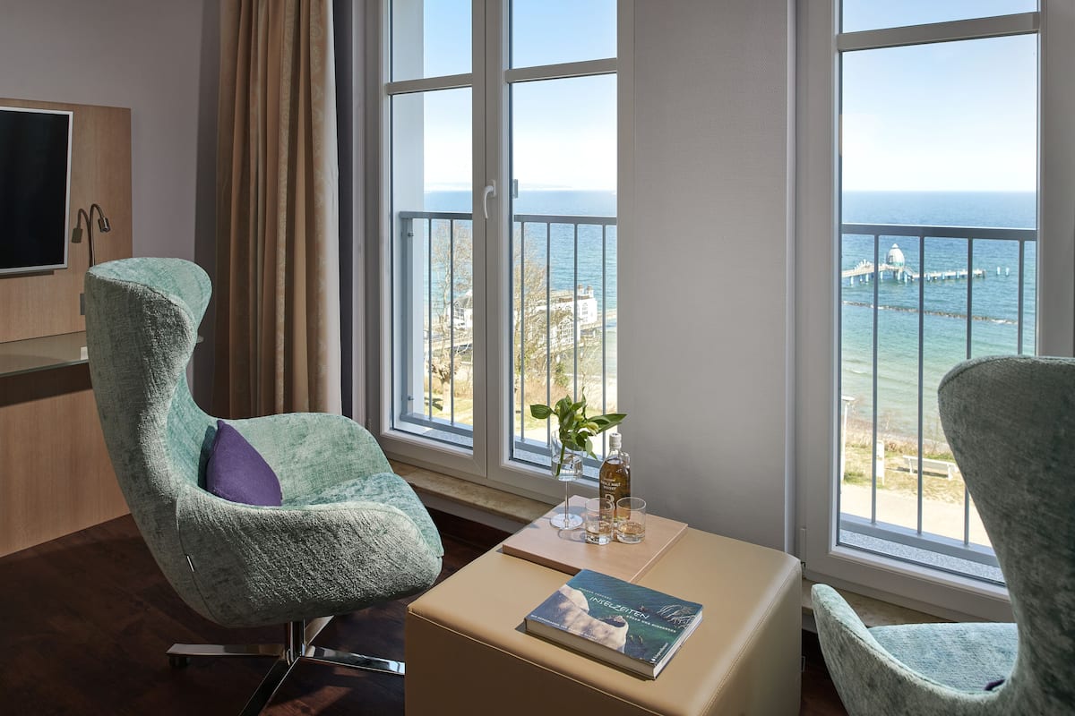 A modern room with a view of the sea, featuring two armchairs, a small table with a book and a bottle, and a TV on the left.