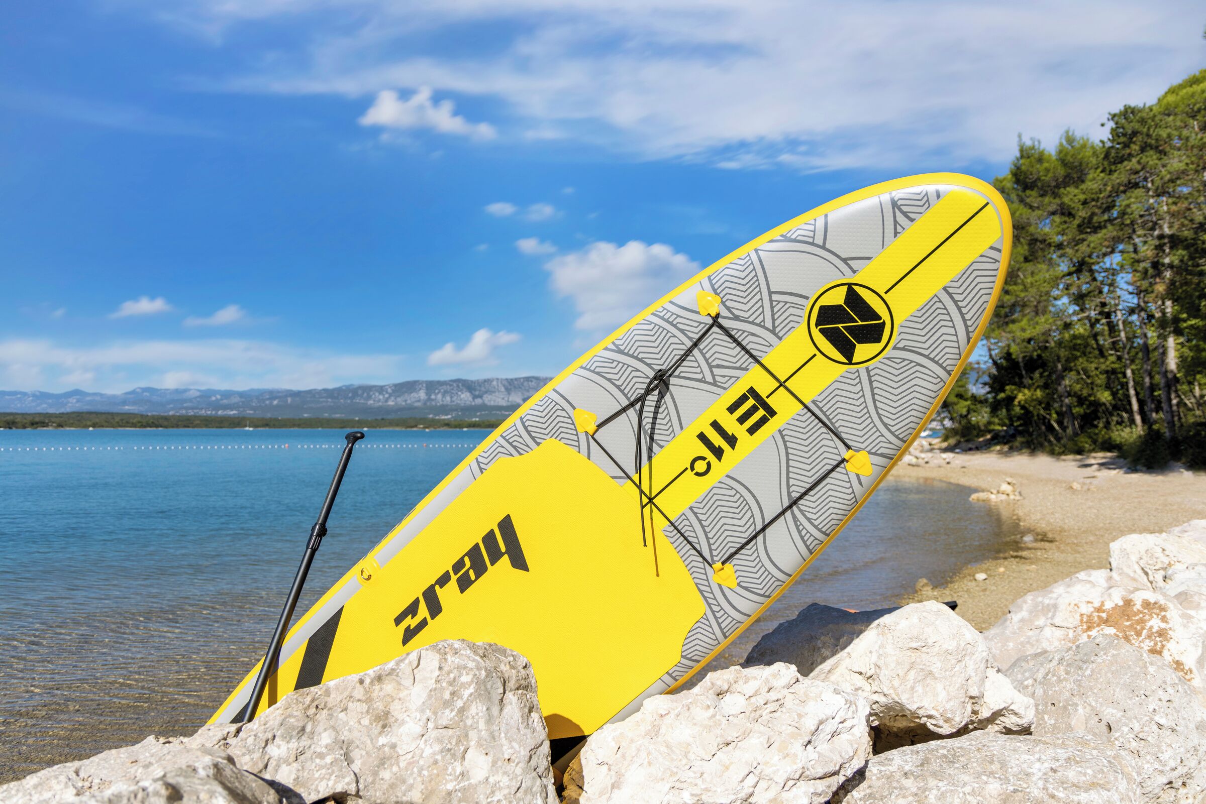 A paddleboard with a yellow and grey design, leaning against rocks by a calm lake with mountains in the background.