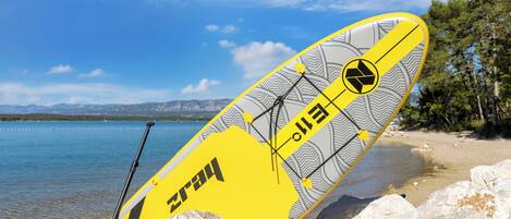 A paddleboard with a yellow and grey design, leaning against rocks by a calm lake with mountains in the background.