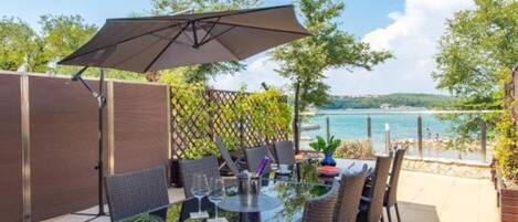 Una zona d'exterior per dinar amb taula i cadires de vidre, un parasol gran i vista al mar.