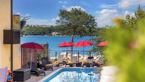 A poolside area with lounge chairs and red umbrellas, overlooking a body of water and a tree. - Soline Bay Seashore Residence (Dobrinj)