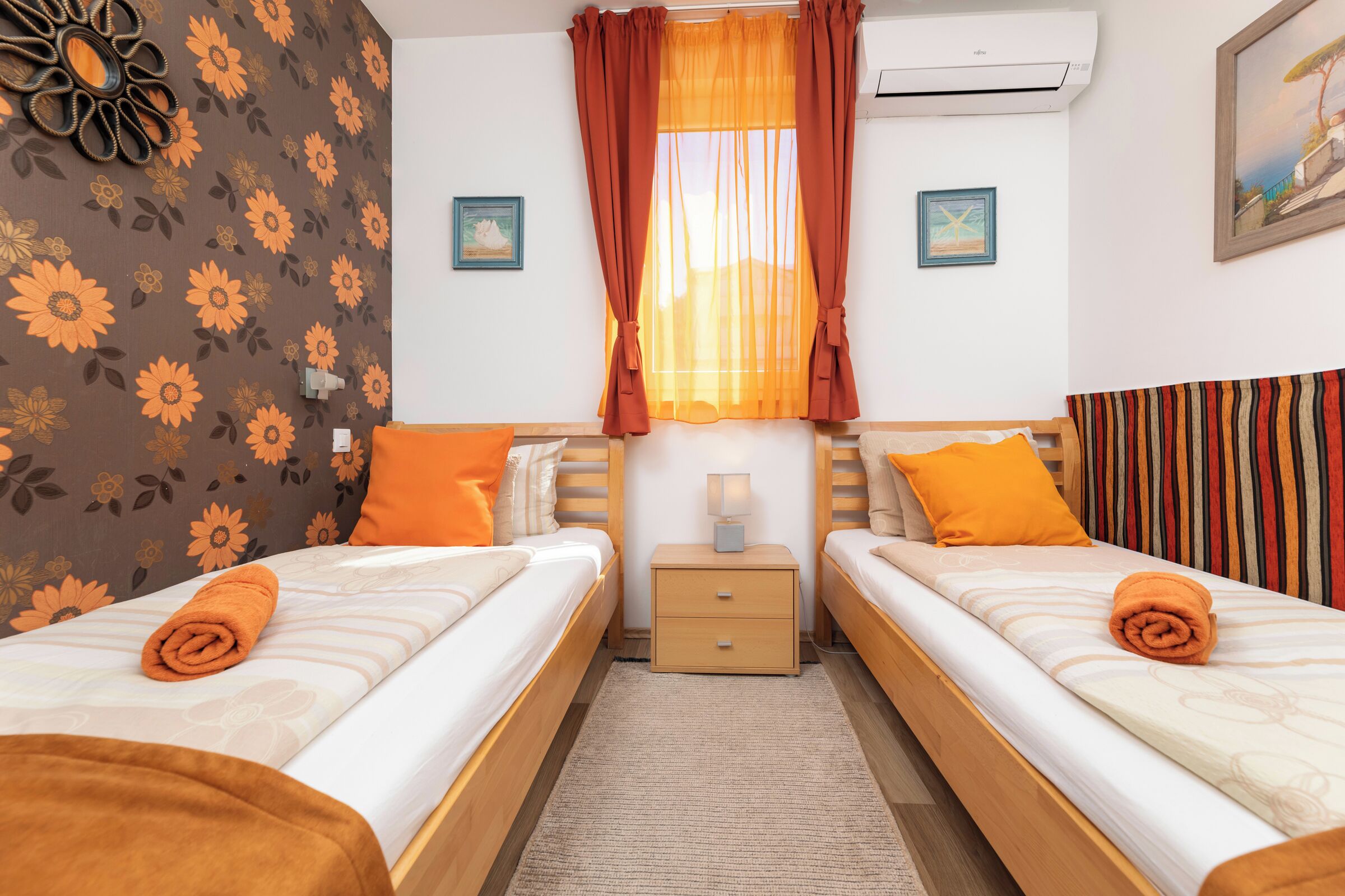 Ein Hotelzimmer mit zwei Betten, einem Nachttisch, einem Fenster mit orangefarbenen Vorhängen und einer Wand mit einem floralen Muster.