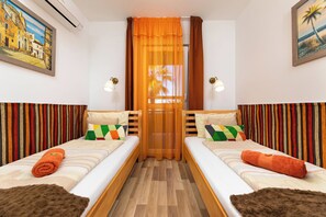 Quarto de hotel com duas camas, piso de madeira e vista para palmeiras através de uma janela com cortinas laranja.