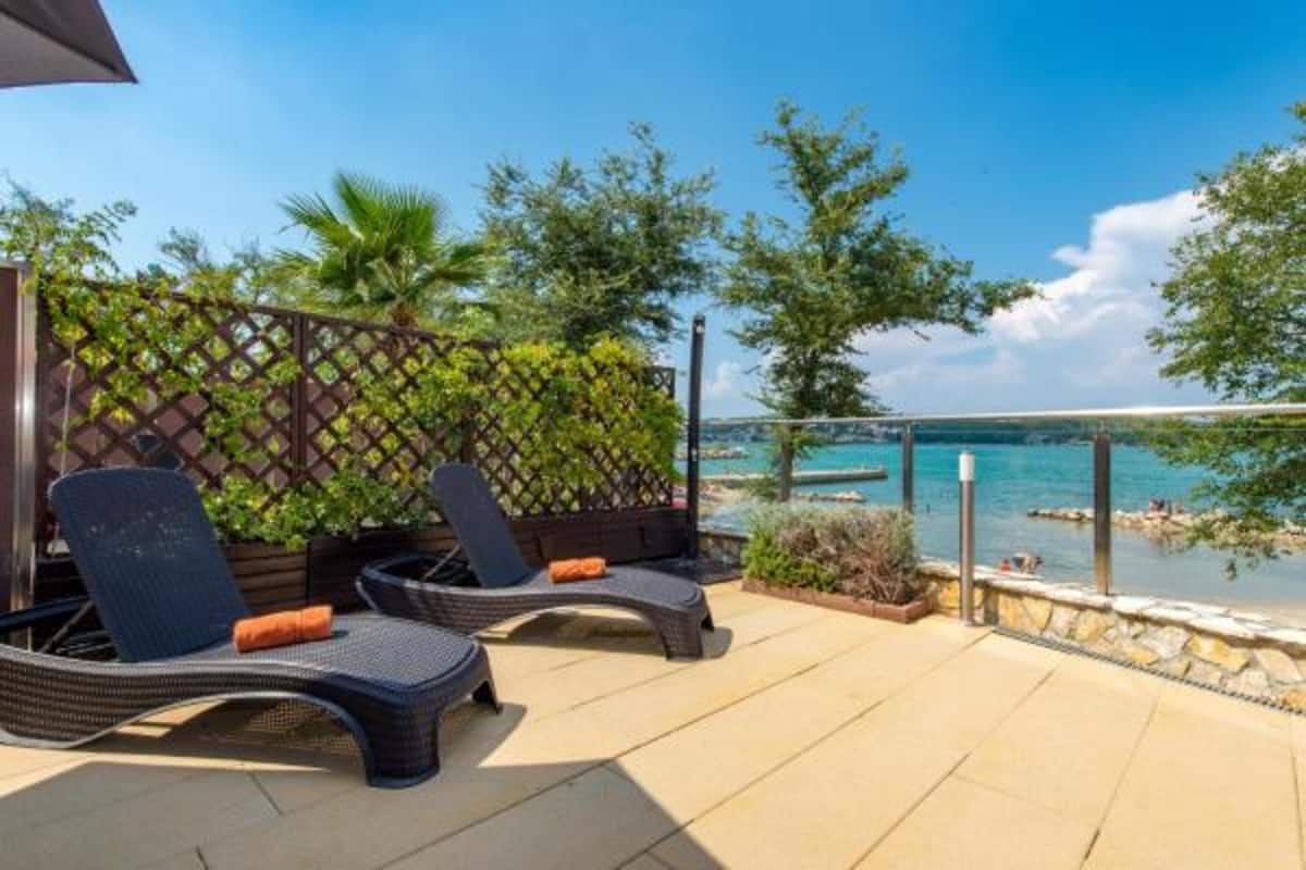 A patio with two lounge chairs and a wooden fence, overlooking a body of water and a beach.