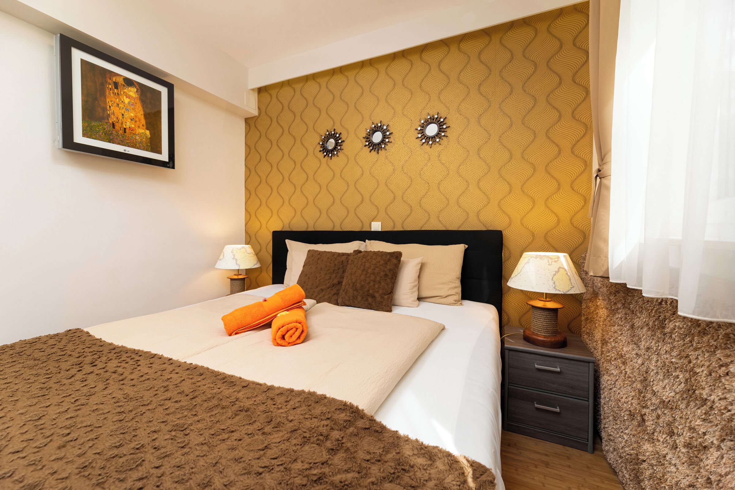 Un letto rifatto con un copriletto marrone, due cuscini e un asciugamano arancione. Sono presenti due comodini con lampade e un quadro appeso al muro.