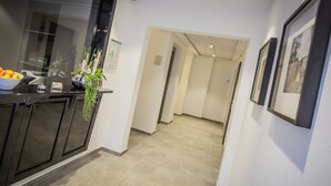 A hallway with a kitchen area on the left, featuring a black countertop, a bowl of fruit, and a hanging plant. There are framed pictures on the right wall and a door at the end of the hallway. - Burghotel Lingen (Lingen)
