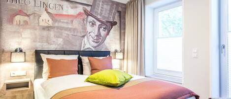 Una habitación de hotel con una cama grande, un cabecero con un mural, una mesita de noche y una ventana con cortinas.