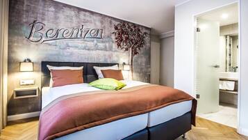Un dormitorio con cama, mesitas de noche, un mural en la pared y un baño visible a través de una puerta abierta.