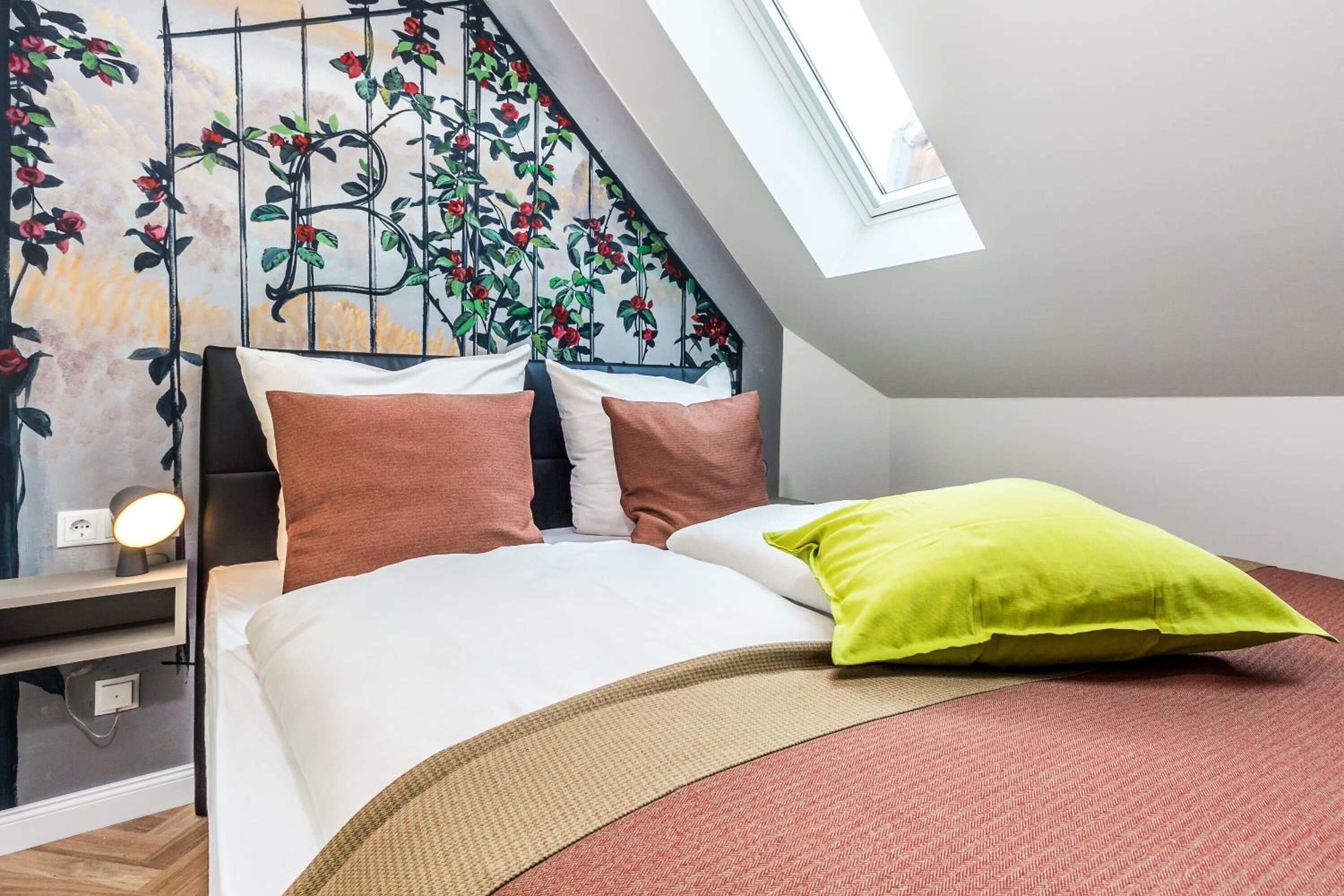 Quarto com cama, almofadas, mesa de cabeceira com abajur, janela e parede com um mural floral.