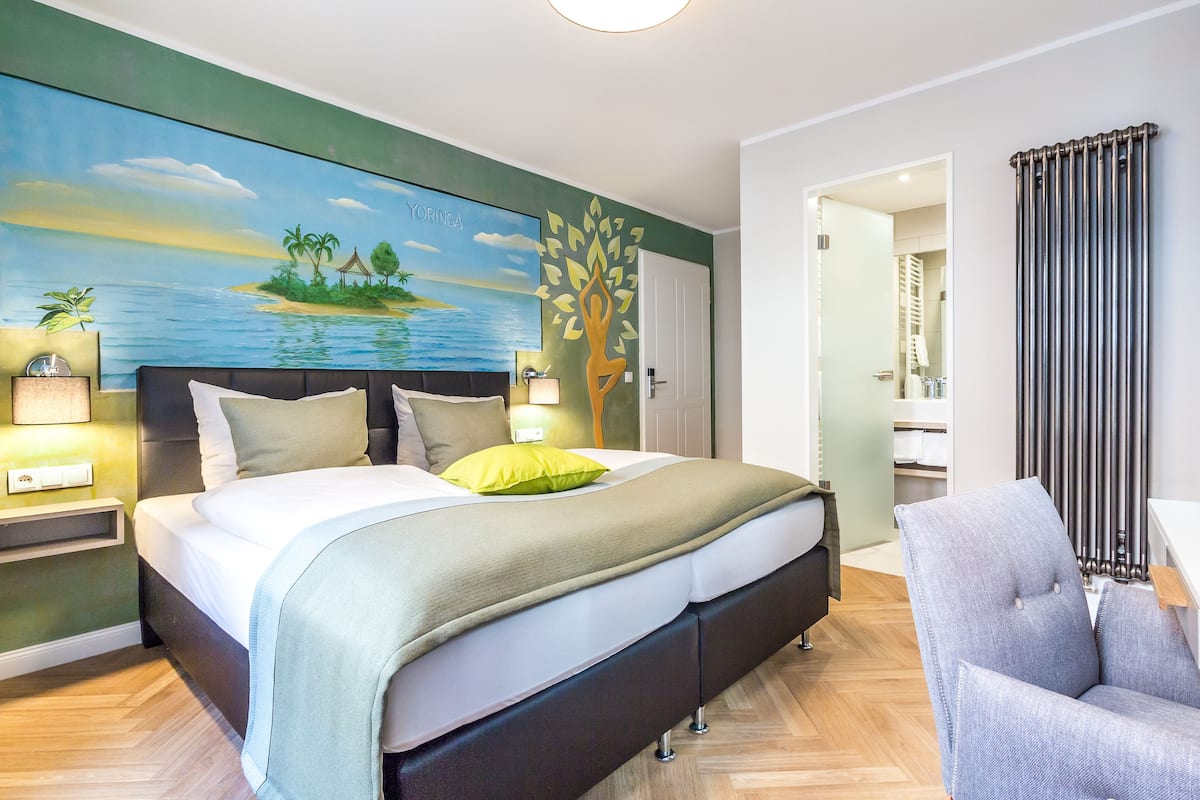 Una habitación de hotel moderna con una cama grande, una mesita de noche, una silla y un cuadro de una escena tropical.