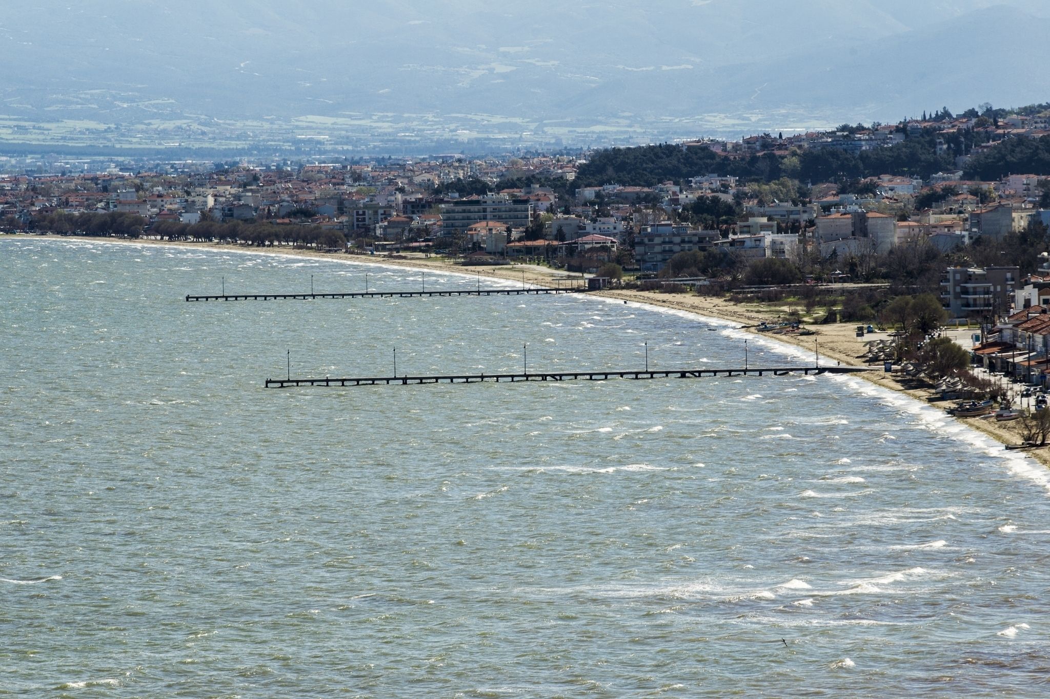 A coastal cityscape with a pier extending into the sea, buildings along the shore, and a hilly background.