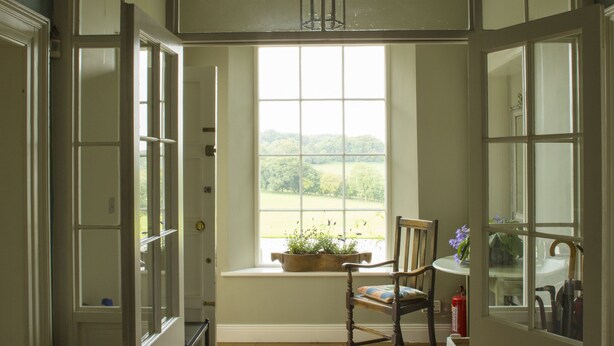 An open doorway leading to a room with a wooden floor, a window with a view of greenery, a chair, and a small table with a plant. - Lough Bawn House (Collinstown)