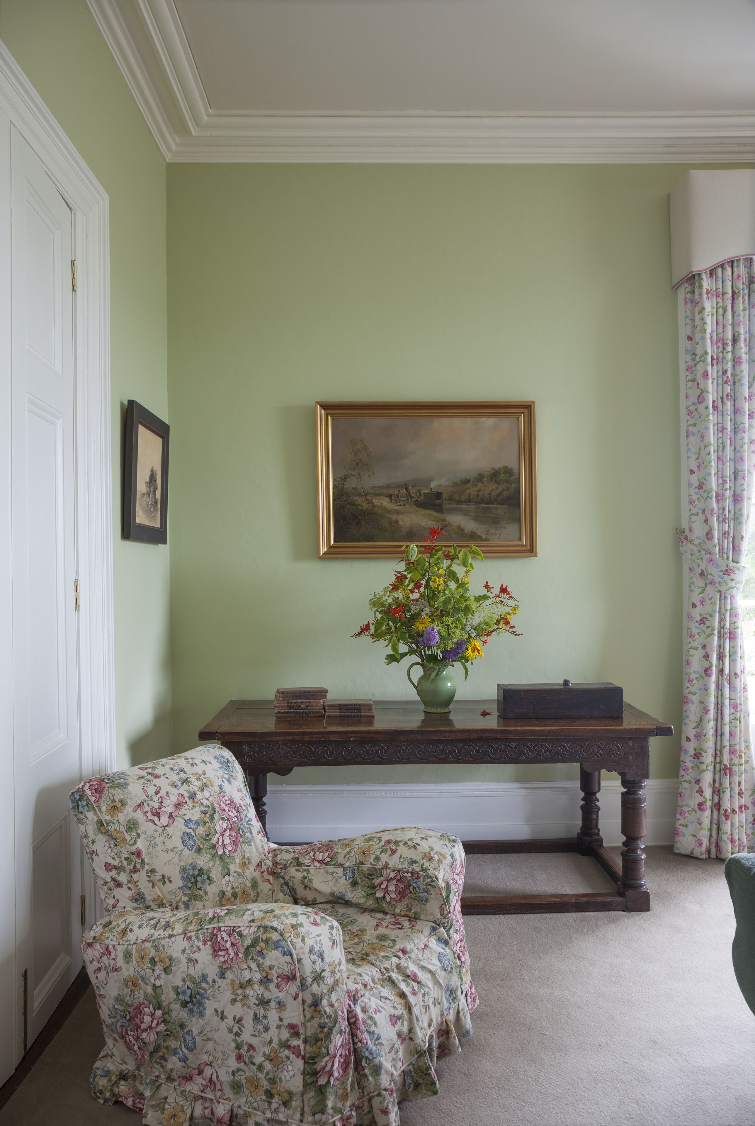Una habitación con una pared verde, un cuadro de paisaje enmarcado, un sillón floral, una consola de madera con un florero y un sillón verde.